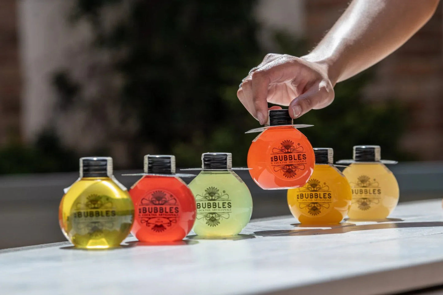 Mano che prende una bottiglia rotonda arancione di cocktail artigianali Mr. Bubbles su tavolo bianco con altre bottiglie colorate