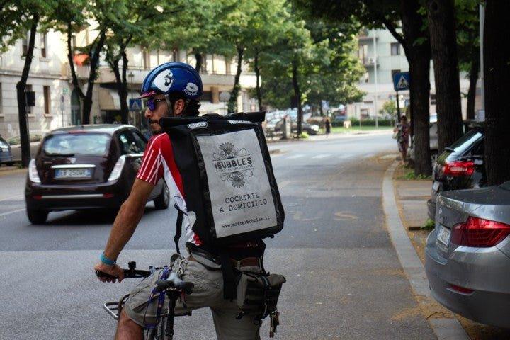 Ciclista con casco blu e maglia rossa consegna cocktail a domicilio in città su strada alberata