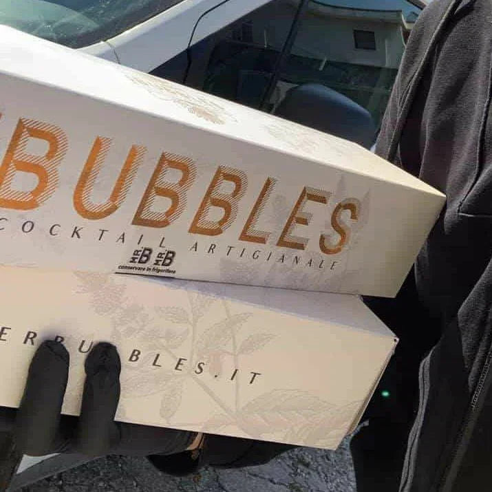 Confezioni di cocktail artigianali Bubbles tenute da una persona con guanti neri, sfondo auto parcheggiata