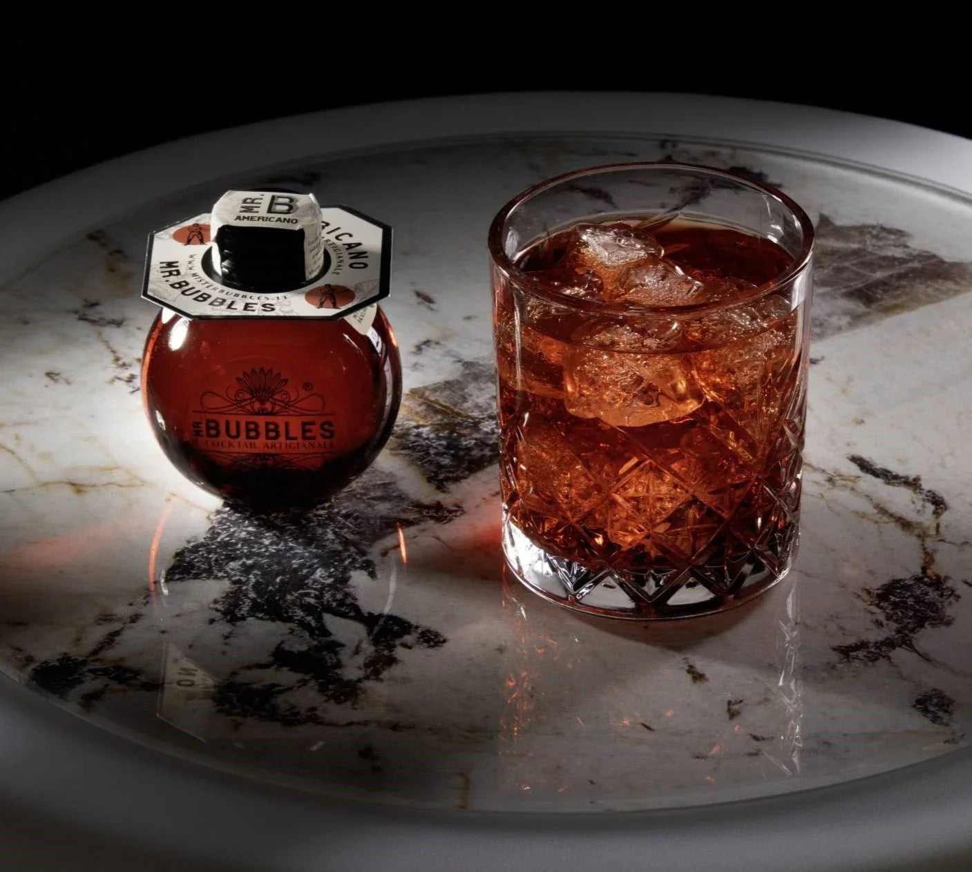 Bicchiere di cocktail Americano con ghiaccio accanto a bottiglia rotonda Mr. Bubbles su tavolo in marmo