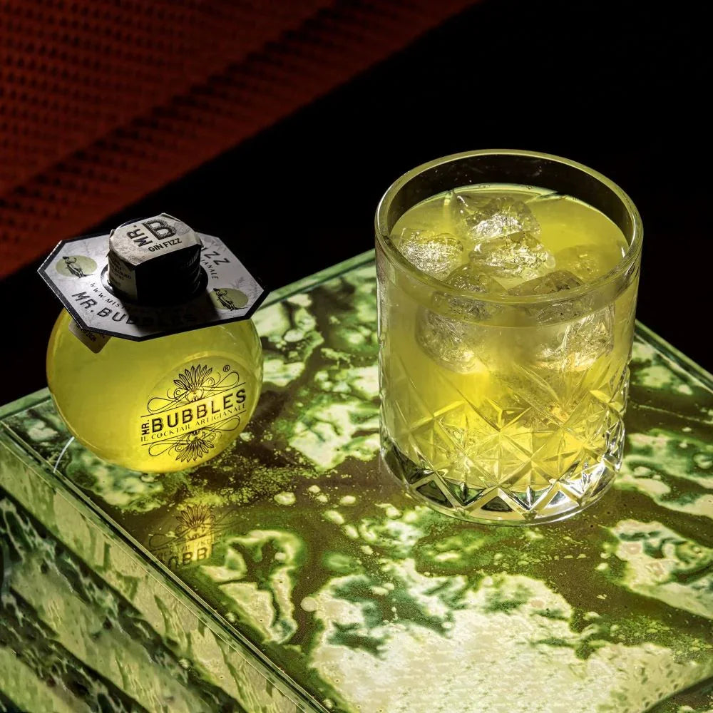 Bottiglia e bicchiere di cocktail Gin Fizz giallo con ghiaccio su tavolo verde marmorizzato