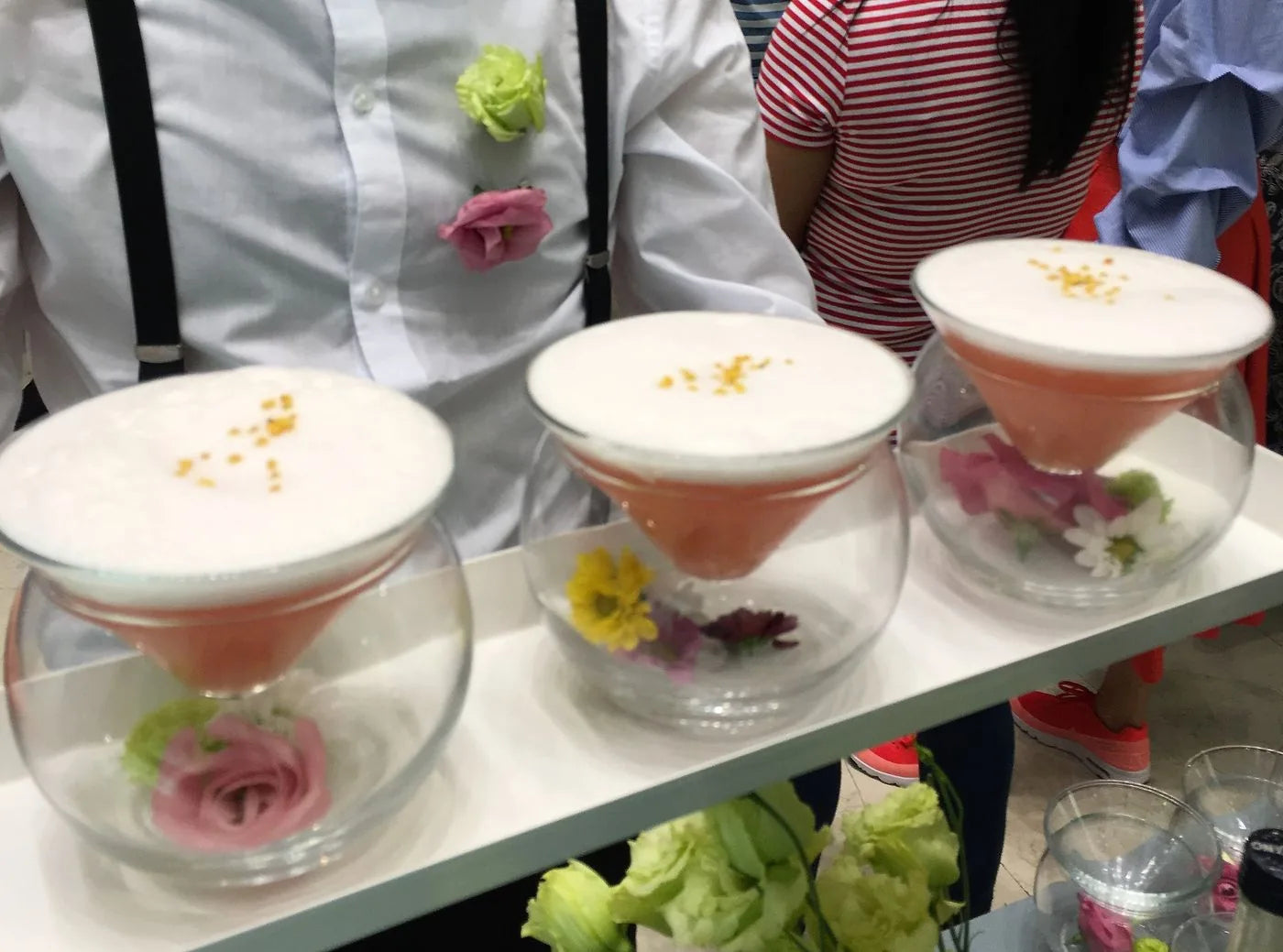 Tre cocktail serviti a un catering: coppette dentro sfere di vetro, fiori commestibili alla base, il barman in camicia e bretelle dietro il vassoio