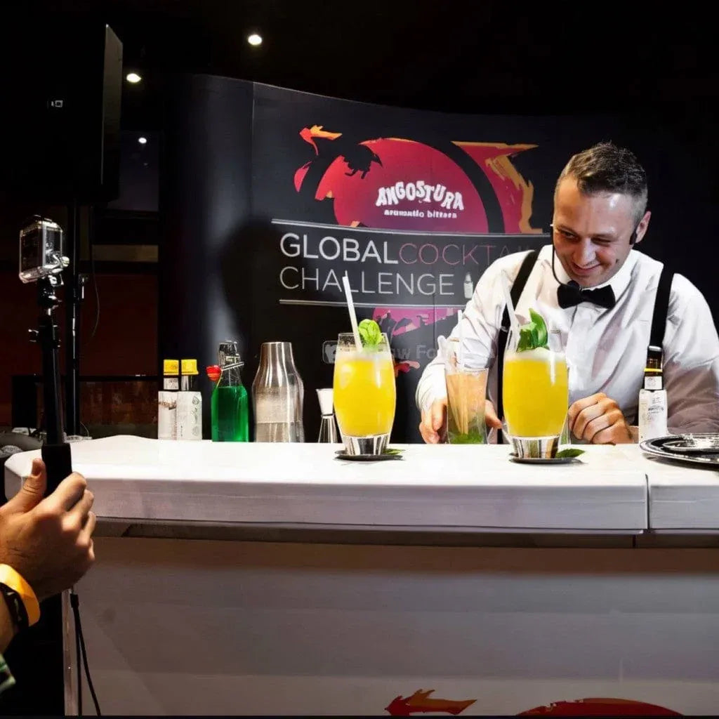 Angostura Global Cocktail Challenge: Matteo in camicia e bretelle appoggia due cocktail gialli sul banco, dietro il fondale della gara