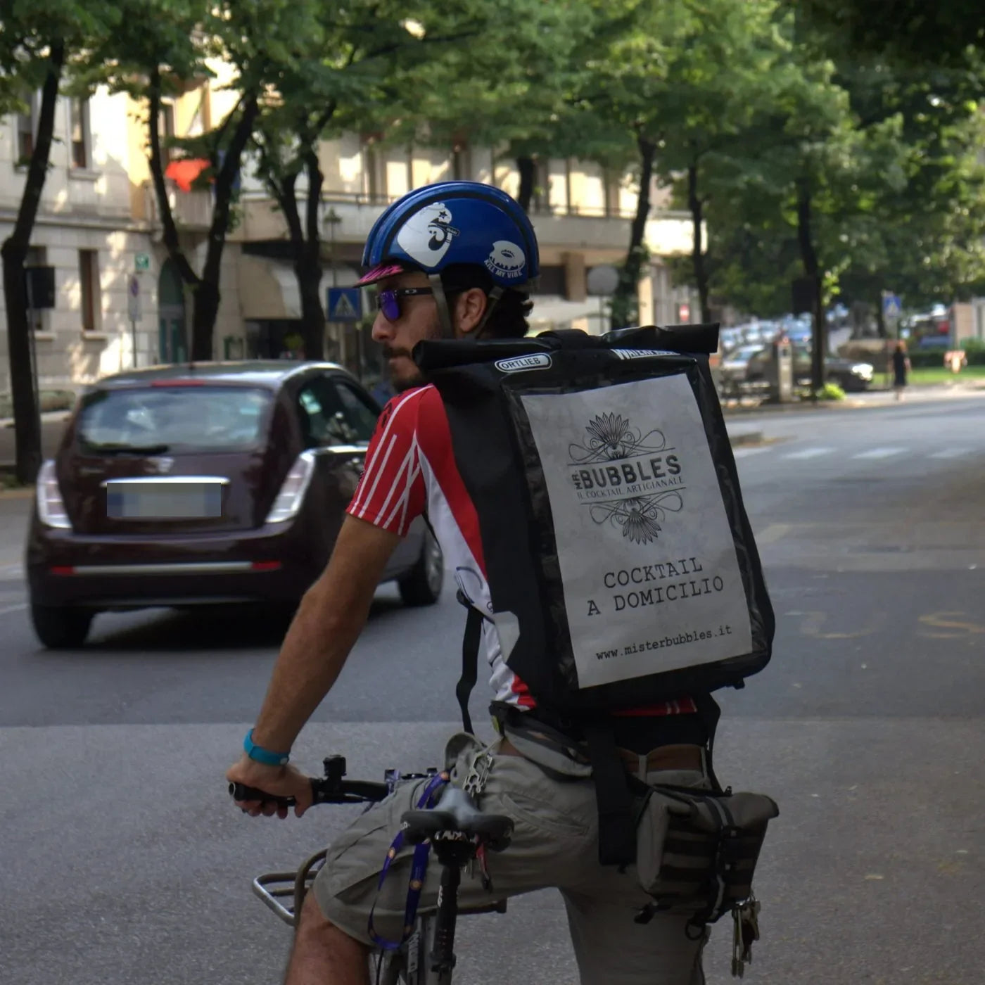 Un corriere in bicicletta di spalle, con lo zaino termico che porta il logo Mr. Bubbles e la scritta cocktail a domicilio
